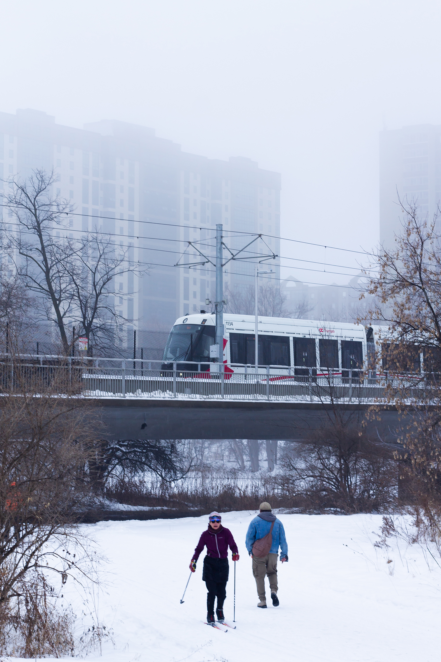 OTrain passing over a bridge while people pass below it on the snow